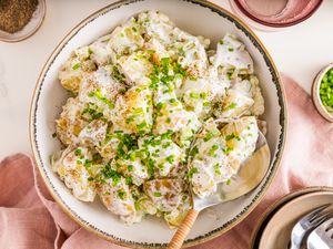 Bowl of Sour Cream and Onion Potato Salad With a Spoon, and in the Surroundings, a Bowl of Seasoning, a Glass of Water, a Bowl of Chives, Stack of Plates With Utensils, and a Blush Table Napkin