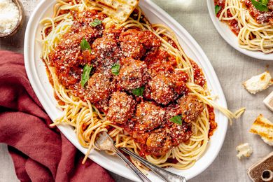 Platter of Spaghetti and Meatballs With a Piece of Toasted Bread and Topped With Fresh Basil Leaves, and in the Surroundings, a Burgundy Table Napkin, a Bowl of Parmesan, Bite Size Bread Pieces Scattered on the Table, and a Plate With More Spaghetti and Meatballs