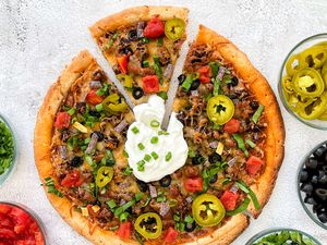 Taco Pizza on the Counter With a Slice Slightly Removed from the Pizza, and in the Surroundings, Bowls of Toppings (L to R: Sliced Scallions, Salsa, Sliced Lettuce, Sliced Olives, and Sliced JalapeÃ±os)