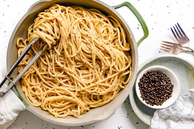 Pot of Vegan Cacio e Pepe with Some Noodles Wrapped around a Pair of Tongs Next to a Bowl of Black Pepper on a Small Plate and Two Forks on the Counter