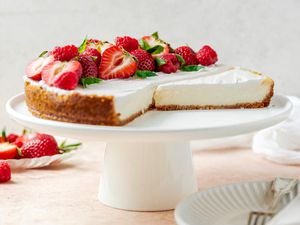 Vegan Cheesecake Topped With Halved Strawberries and Raspberries on a Cake Platter, and on the Counter Surrounding It, a Small Plate With More Fruit and Another Small Plate With a Slice of Cheesecake 