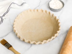 Vegan Pie Crust (Not Baked) in a Pie Dish, and Surrounding It, a Small Bowl of Flour, a Rolling Pin, a Pastry Brush, and a Kitchen Towel