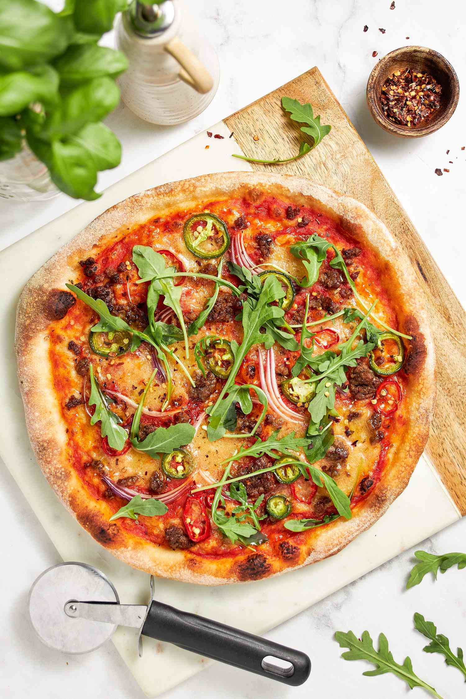 Vegan Pizza Topped With Jalapeno and Red Onion Slices, Arugula, and Chili Flakes on a Cutting Board With a Pizza Cutter, and in the Surroundings, a Glass With Fresh Basil, a Oil Dispenser, a Bowl of Chili Flakes, and a Few Rogue Arugula on the Counter