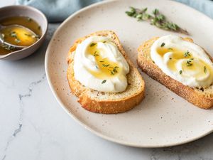 Two Pieces of Whipped Ricotta Toast Topped with Lemon-Thyme Honey Next to a Bowl of More Honey