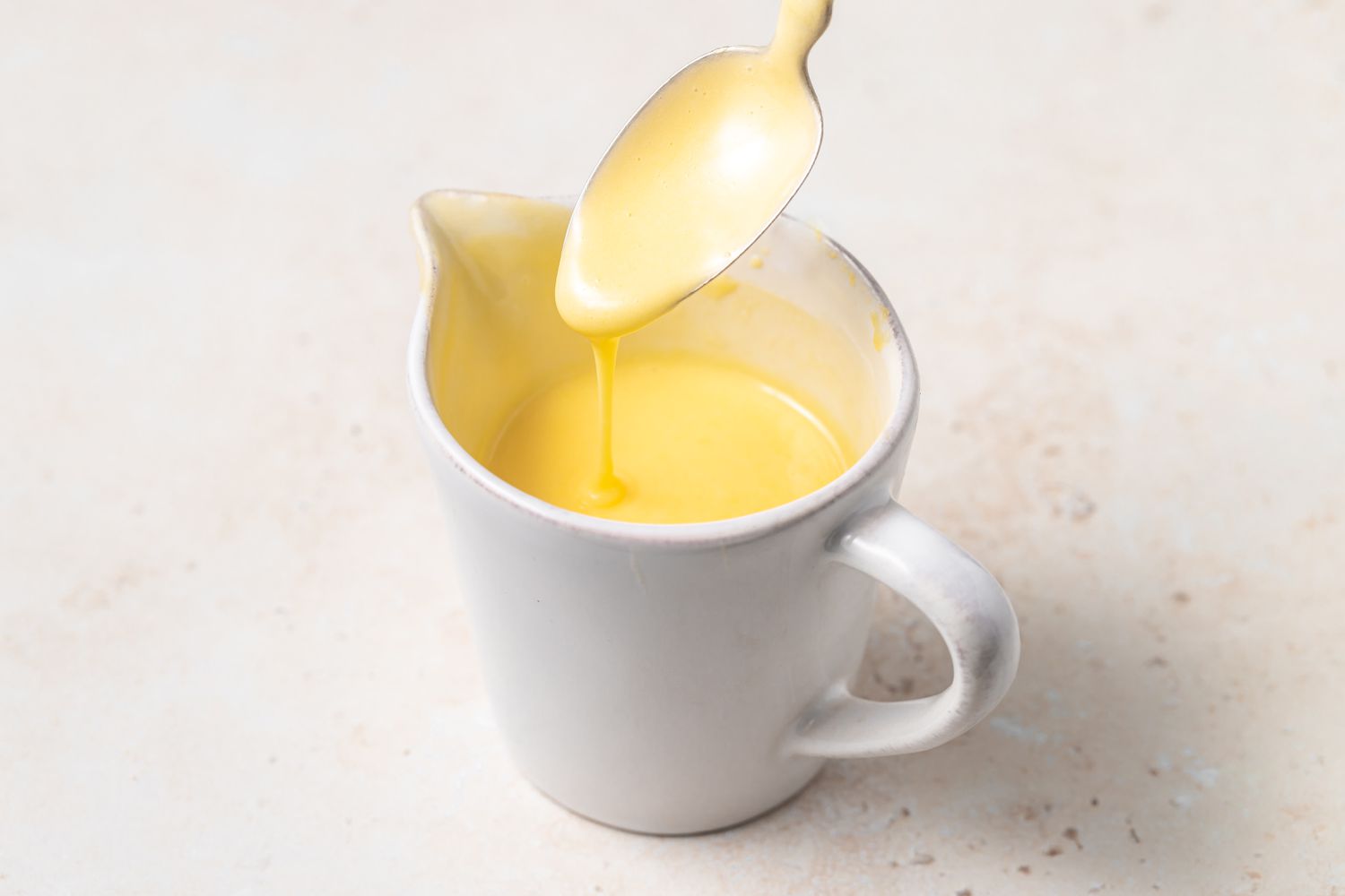 Blender Hollandaise Sauce in a spouted cup with a spoon letting the sauce drip back into the dish.