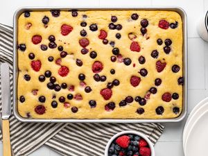 Sheet Pan Buttermilk Pancakes with Blueberries and Raspberries Surrounded by a Stack of Plates, a Cup of Coffee, a Bowl of Berries, and Serving Utensils on a Kitchen Towel