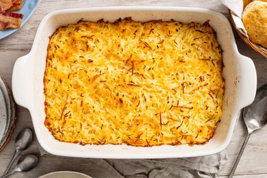 Copycat Cracker Barrel hash brown casserole in a casserole dish at a table setting with a plate, a basketful of bisquits, a stack of plates, and spoons on the counter 