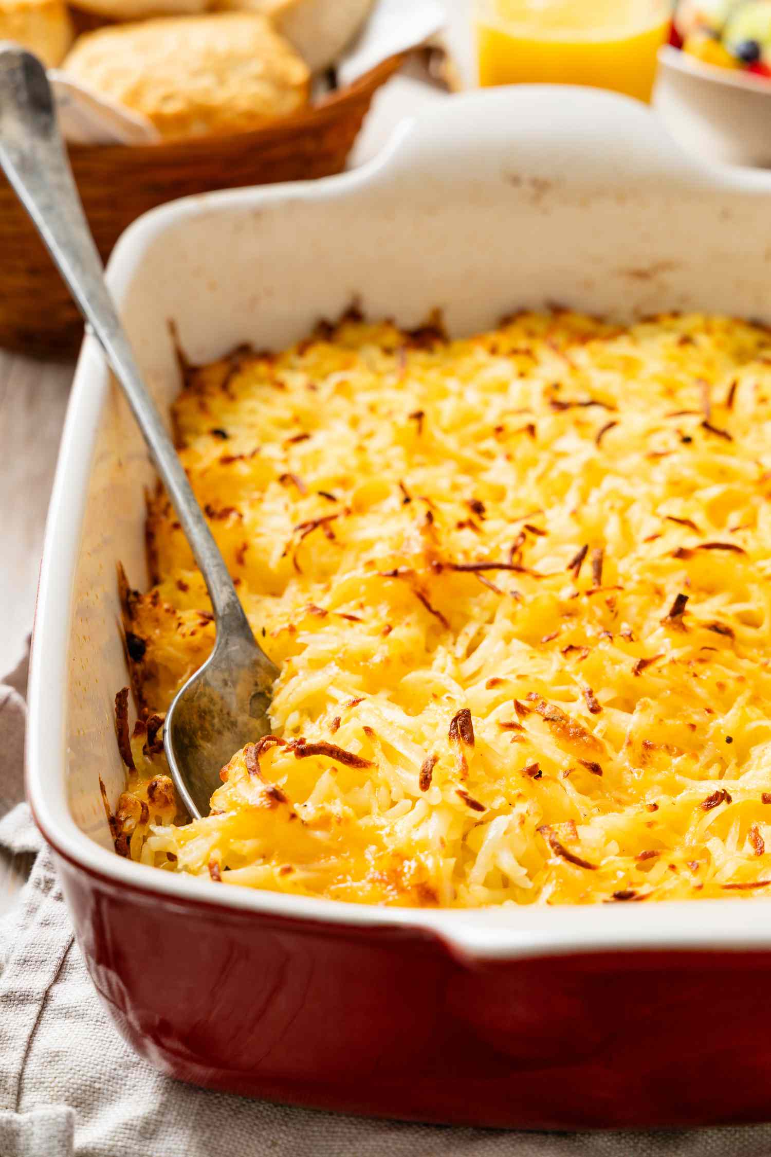 Spoon digging into copycat Cracker Barrel hash brown casserole in a casserole dish with a basketful of biscuits and a glass of orange juice in the background