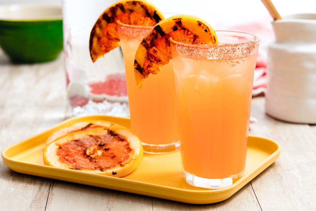 Two Salt Rimmed Glasses of Grilled Grapefruit Paloma With Two Grilled Grapefruit Slices on the Glass Rim Next to More Slices of Grilled Grapefruit on a Small Tray, and in the Surroundings, a Bottle of Club Soda and a Cream Pitcher