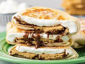 Cross-Section Cut of a S'mores Crunchwrap on a Plate, and in the Background, a Bowl of Mini Marshmallows and a Plate With a Stack of Graham Crackers
