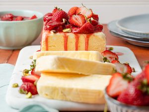 Strawberries and Cream Semifreddo Slices Cascading on a Platter Next the Rest of the Uncut Semifreddo on the Same Platter. Semifreddo Is Topped With Halved Fresh Strawberries and Strawberry Compote. In the Surroundings, a Bowl of Fresh Strawberries and a Stack of Plates.