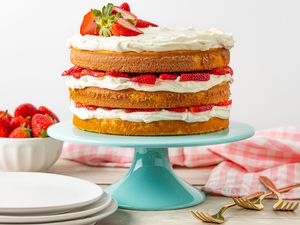 Strawberry Shortcake Cake on a Cake Stand, and in the Surroundings, a Stack of Plates, a Bowl With Whole Strawberries, a Kitchen Towel, and a Few Forks on the Counter