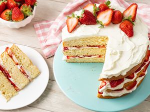 Strawberry Shortcake Cake Topped With Halved Strawberries on a Cake Stand, and on the Counter Next to It, a Plate With a Slice of Cake, a Bowl of Strawberries, and a Kitchen Towel