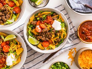 Bowl of Taco Salad Surrounded by Two More Bowls With Servings of Taco Salad, a Bowl of Salsa, a Bowl of Quartered Limes, a Bowl of Cheese, a Bowl of Sliced Green Onions, and a Bowl With Utensils, All on a Kitchen Towel